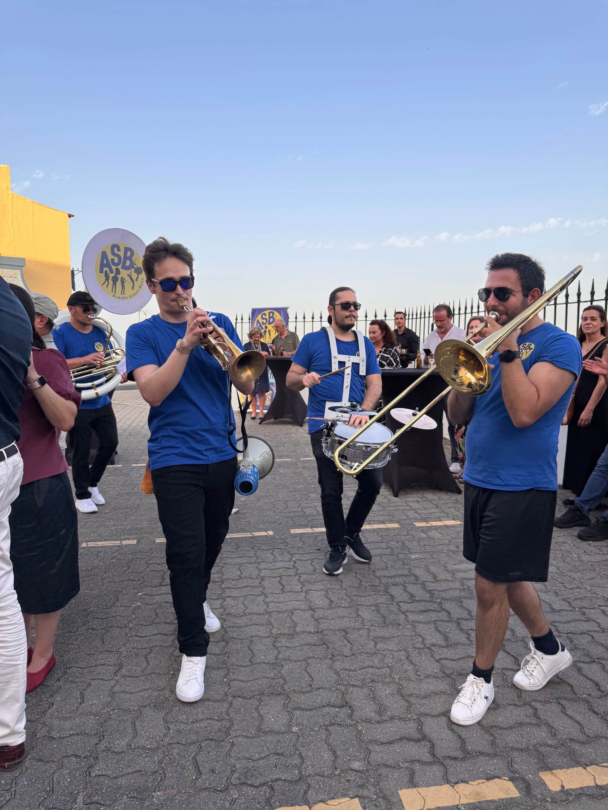 A banda a desfilar por uma calçada, com trompete e trombone, e o público em volta