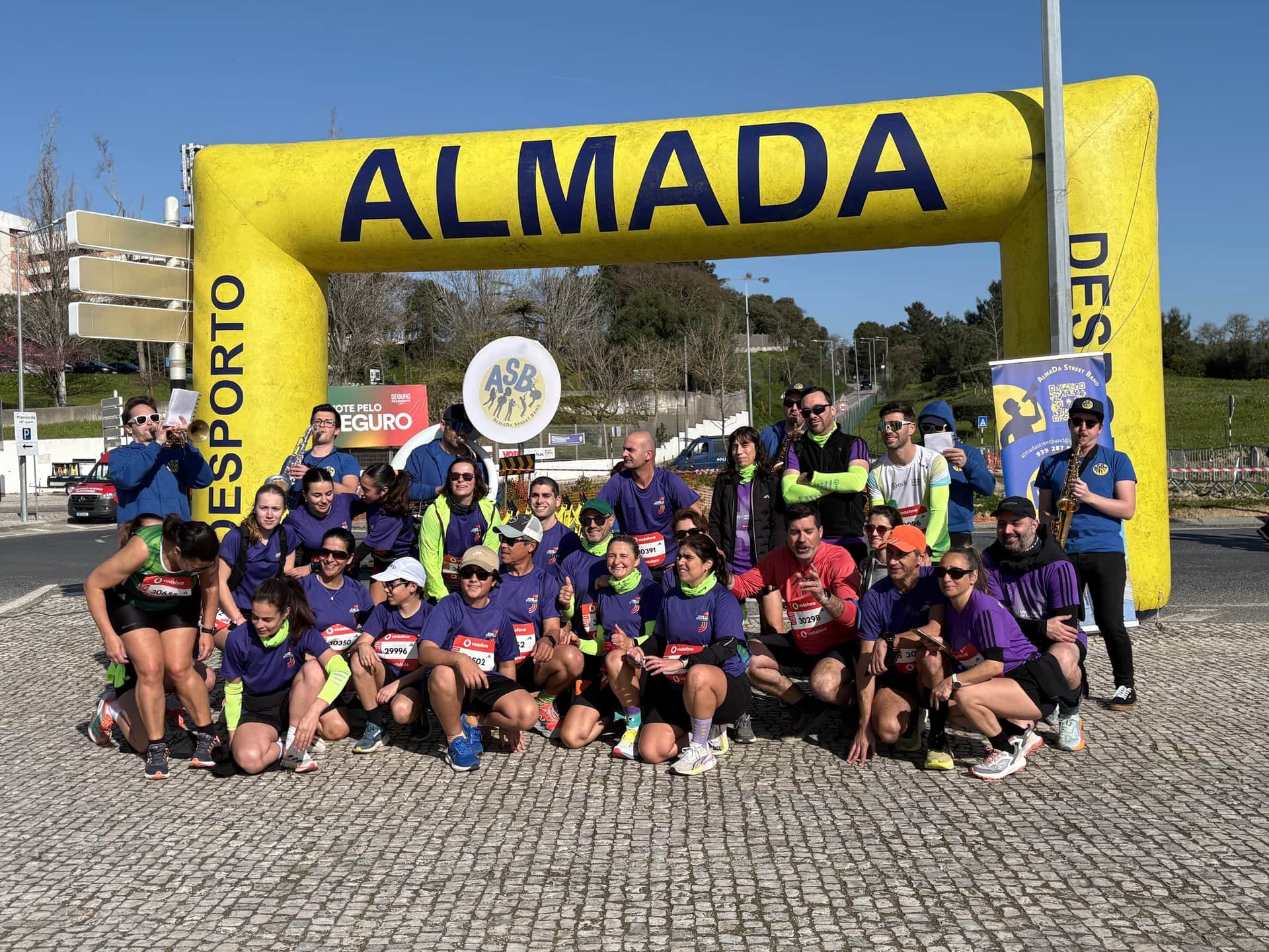 A banda e um grupo de participantes sob o pórtico amarelo de Almada Desporto