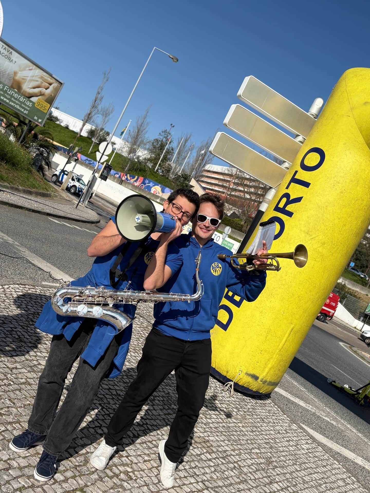 Dois músicos com megafones e saxofone junto ao pórtico amarelo