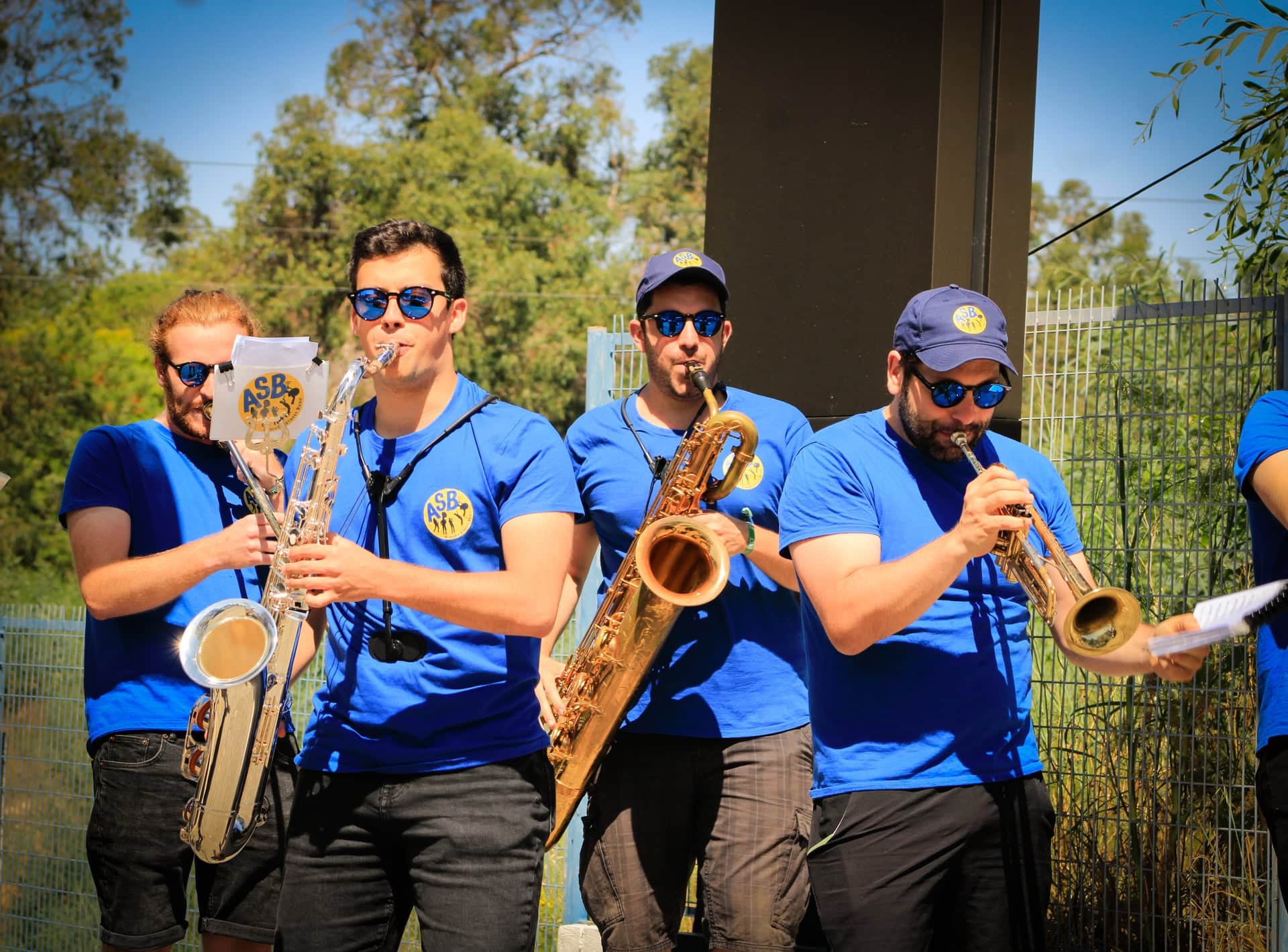 Saxofonistas e trompetista a tocar ao sol junto a um jardim