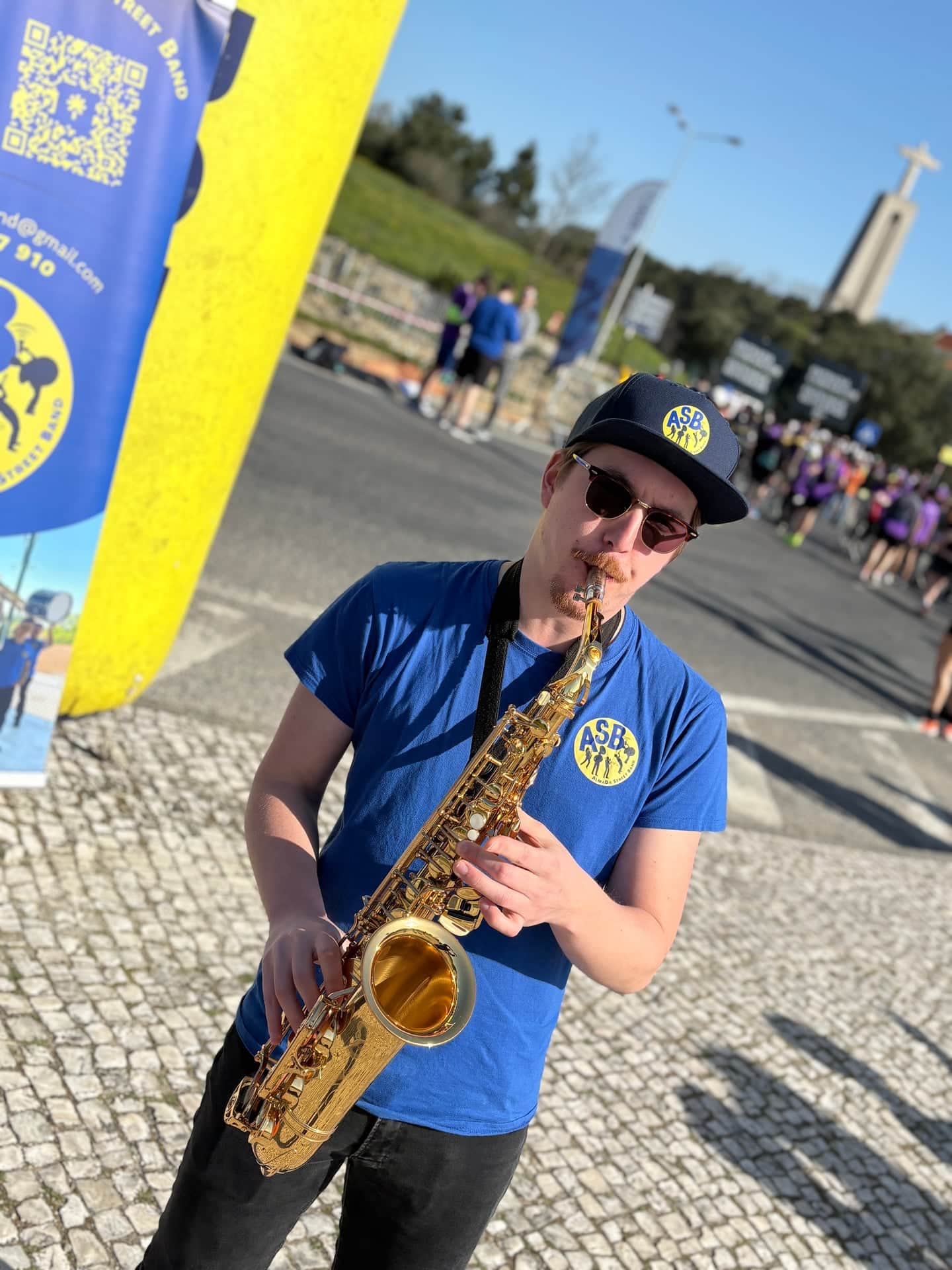 Saxofonista de boné preto e óculos escuros junto à bandeira da ASB